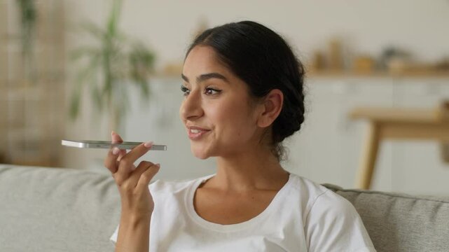 Happy smiling Indian woman at home Arabian positive Arabian girl female lady at living room mobile phone talking microphone smartphone mic talk loudspeaker record voice mail recording audio message