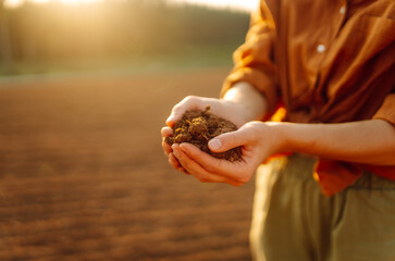 A young farmer kneels on the ground at sunset, cradling a handful of dark, fertile soil while...