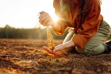 A young farmer kneels on the ground at sunset, cradling a handful of dark, fertile soil while...