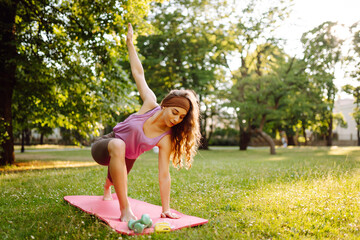 A woman practices yoga in a sunlit park while using a mat and weights on a warm afternoon. . Healthy lifestyle, sport. Fitness.