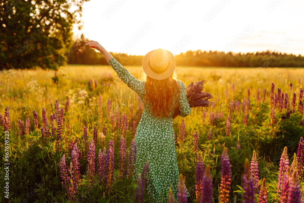 Wall mural Woman in a green dress enjoys a sunny evening in a field of colorful lupins while holding a bouquet in the golden hour sunlight. Collection of medicinal herbs.