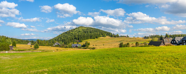 The tranquil Jizerka village rests at the foothills of Bukovec Mountain, surrounded by lush greenery and under a bright, open sky. The idyllic landscape showcases the charm of the Jizera Mountains.