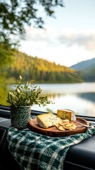 Scenic picnic setup with cheese, crackers, and greenery by a tranquil lake surrounded by mountains, perfect for outdoor relaxation.