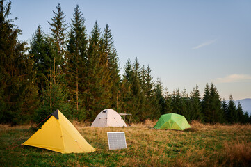 Solar panel in grassy field with tents in background highlights eco-friendly camping setup, surrounded by dense evergreen trees in evening. Integration of renewable energy in outdoor activities.