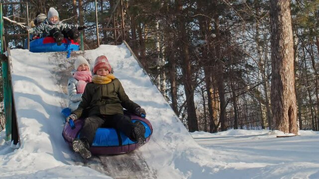 Slowmo shot of diverse little kids in warm clothes sliding down icy slide on inflatable sledge snow tubes in winter park on sunny day