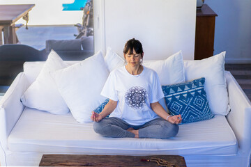 Mid-adult woman meditating on her sofa at home, with natural daylight and palms pressed together at...
