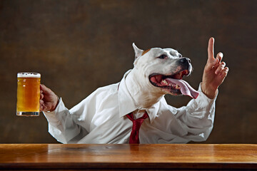 White dog wearing shirt and tie, sitting at desk, holding mug of beer, and raisin ginger upwards symbolizing brilliant ideas come in relaxing atmosphere. Concept of animal themes, fun, business