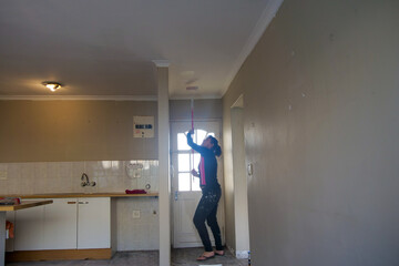 Woman standing and painting a ceiling with an extended roller and looking up at the ceiling. She is...