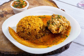 A vibrant, traditional Ecuadorian dish, Seco de Pollo, served with flavorful yellow rice and fried plantains. The dish features a tender chicken leg simmered in a rich, aromatic sauce made from tomato