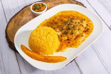 A vibrant, traditional Ecuadorian dish, Seco de Pollo, served with flavorful yellow rice and fried plantains. The dish features a tender chicken leg simmered in a rich, aromatic sauce made from tomato