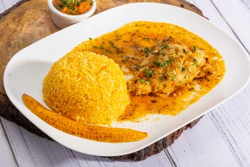 A vibrant, traditional Ecuadorian dish, Seco de Pollo, served with flavorful yellow rice and fried plantains. The dish features a tender chicken leg simmered in a rich, aromatic sauce made from tomato