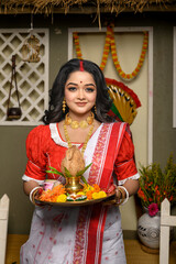 Portrait of young woman, adorned in traditional saree, complemented by gold jewellery, holding a plate filled with religious offerings. Indian culture, festivals and fashion.