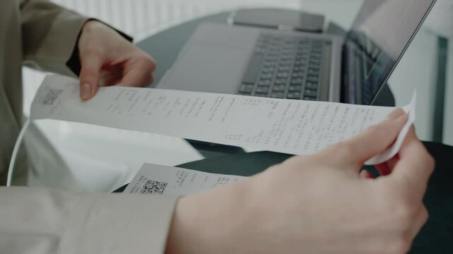 A close-up image of hands holding receipts next to a laptop, depicting business accounting or personal finance management in an office or home setting. Hands Examining Receipts While Working