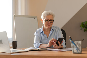 Confident stylish european middle aged senior woman using smartphone at workplace. Stylish older mature 60s gray haired lady businesswoman with cell phone in office. Boss leader using internet apps