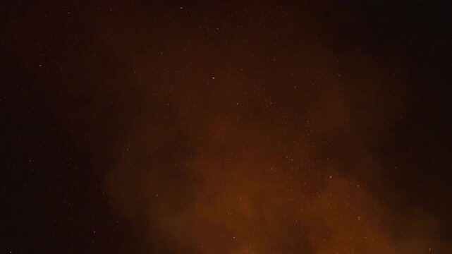 Fire sparks and ember particles from a bonfire light up the dark night sky as they rise up with smoke