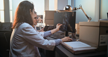 Two diverse scientists discuss 3D visualization of ancient human head anatomy displayed on PC and tablet computer screen, conduct historical research. Archaeologists work in archaeological laboratory.