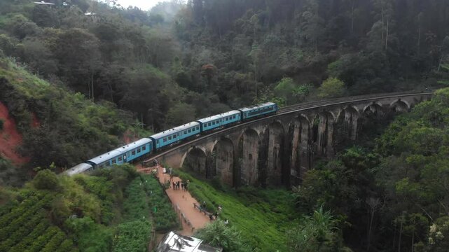 Aerial view of the nine arches bridge with a train passing through lush tea fields and forest under a cloudy sky, Ella, Sri Lanka.