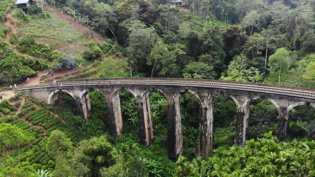 Aerial view of nine arches bridge over railway surrounded by tea fields and forest under a cloudy sky, Ella, Sri Lanka.