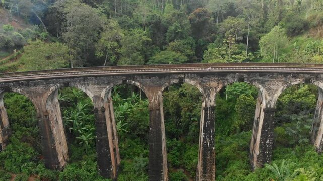 Aerial view of nine arches bridge with a train passing through lush tea fields and a cloudy sky, Ella, Sri Lanka.