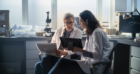 Two diverse scientists in lab coats discuss scientific research, look at papers and use tablet...
