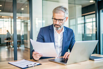 Fototapeta premium Professional Man Working on Laptop in a Modern Office, Holding Documents in a Productive Setting