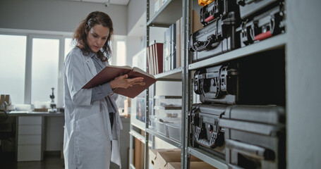 Female scientist or lab worker stands near shelf with folders, protective cases and boxes with fossils, reads scientific book and searches information for research. Modern archaeological laboratory.