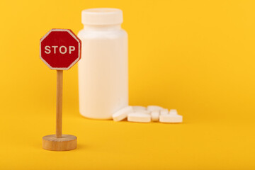 White pill bottle with scattered pills and a wooden stop sign on a vibrant yellow background