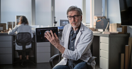 Mature scientist speaks to camera, shows 3D model of human head on tablet computer during interview...