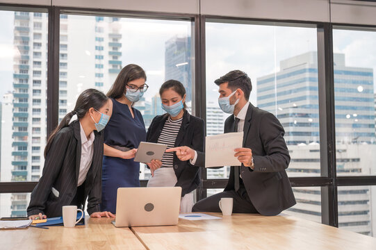 Multiethnic business team wearing face mask during business meeting in modern office. Diverse corporate colleagues brainstorming with technology device