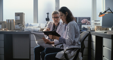 Male and female scientists in lab coats discuss scientific research, look at papers and use tablet...