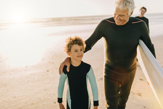 Grandfather and grandson in wetsuits walking on the beach with surfboards