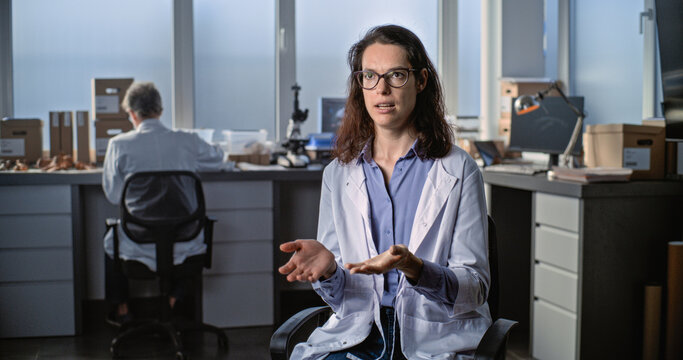 Female young scientist in lab coat talks to the camera and gives interview for popular science film while sitting in archaeological scientific lab. Male colleague works in the background. Static Shot.