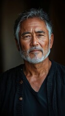 An elderly man with a grey beard and short hair stands in a black shirt with a thoughtful expression on his face.