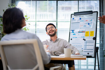 A man is talking to two other people in a meeting room. The man is wearing glasses and is looking at a white board with a lot of information on it