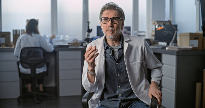 Mature scientist in lab coat speaks to the camera and gives interview for popular science film sitting in archaeological scientific laboratory. Female colleague works in the background. Static Shot.