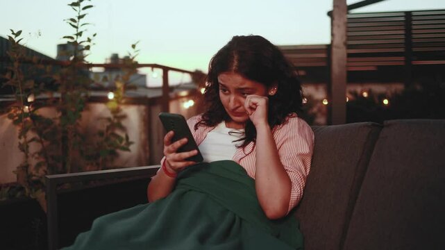 Portrait of crying young indian woman with smartphone sitting on the couch under the blanket and reading bad news email break up message from ex-boyfriend or personal problems at home terrace late