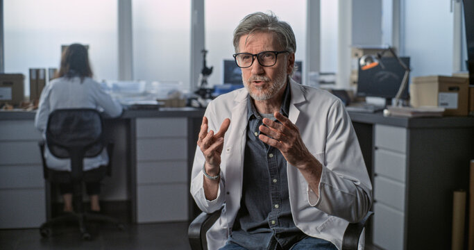 Mature scientist in lab coat speaks to the camera and gives interview for popular science film sitting in archaeological scientific laboratory. Female colleague works in the background. Static Shot.