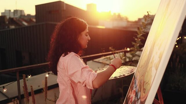 Portrait of focused young indian woman artist painting picture on canvas creating artwork at sunset in creative workshop terrace Stylish lady female enjoying leisure time with art hobby indoors