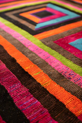 This colorful handwoven textile features a geometric pattern with vibrant shades of pink, green, orange, brown, black, and blue, blending modern and traditional artistic styles