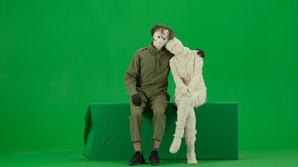 Fototapeta premium Portrait of person in a mummy bandage on chroma key green screen background. Mummy and maniac sitting together hugging looking at the camera.
