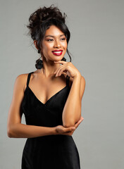Fashion Asian Woman in Black Elegant Dress. Beautiful Lady with Wedding Hair Updo over Gray isolated. Happy smiling Girl in Cocktail Gown with Red Lipstick Makeup and Stylish Hairstyle