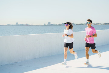 A couple jogs along a sunlit waterfront promenade, enjoying a bright, peaceful morning workout.