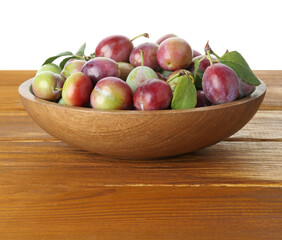 Fresh plums and green leaves in bowl on wooden table against white background