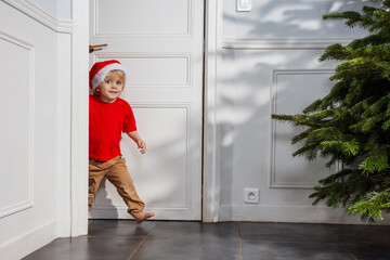 Little boy in red shirt and Santa hat peek out from behind door
