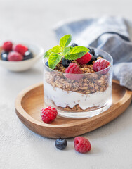 Greek yogurt with muesli or granola, raspberries and blueberries in a glass on a wooden board on a light background