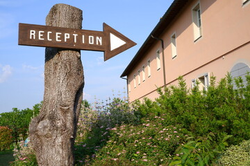 A wooden reception sign mounted on a tree points towards a building, surrounded by greenery and flowers in a rural setting.
