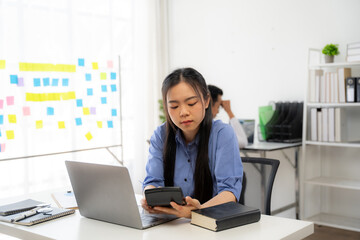 Financial Focus Amidst Office Hustle: A young Asian professional intently calculates expenses, her...