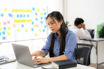 Focused on the Task:  A young businesswoman diligently works at her laptop in a modern office environment, showcasing the dedication and concentration required for success. The blurred figure in the b