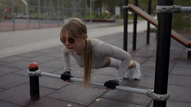 A focused young woman in sunglasses trains on parallel bars at a park, demonstrating determination and fitness. Young Woman Exercising on Outdoor Parallel Bars in the Park
