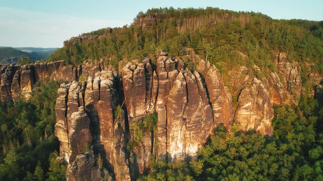 A towering sandstone rock formation rises above a forested area, illuminated by the soft light of the setting sun, showing weathered rock textures.